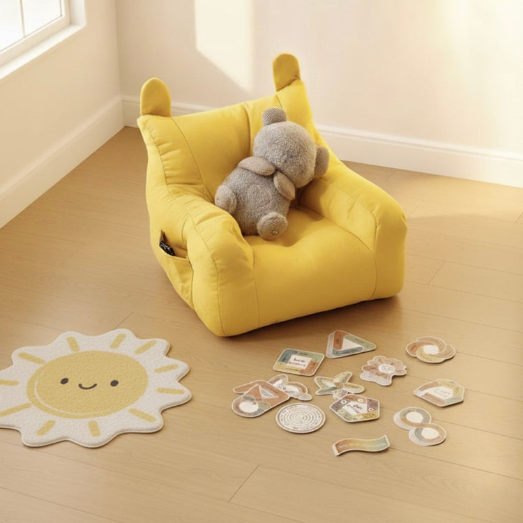 A serene kids' calm corner with a yellow bear-eared beanbag couch, a plush grey teddy bear breathing buddy, and mindful breathing stickers on a light wooden floor. A round, subtly patterned mat completes this inviting relaxation space for children.