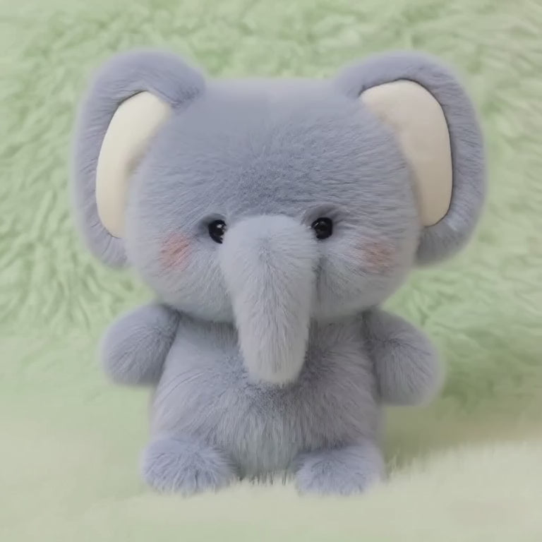  A short, static video of a fluffy, blue-grey plush elephant. It has large ears, a long trunk, and rosy cheeks, and it sits in an upright, empowered posture on a soft, mint-green fluffy surface.