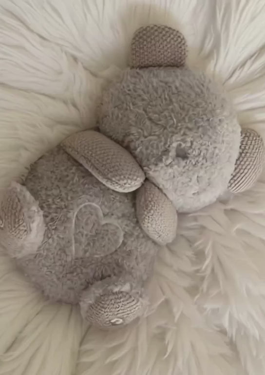 A short video showing a grey plush toy, called a Breathing Buddy, lying face down on a white faux fur rug. The toy's back gently rises and falls in a rhythmic motion that mimics breathing, while a soft, heart-shaped light on its back fades in and out in time with the movement.