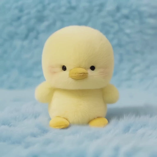 A short, static video of a fluffy, yellow chick plush toy. It has a round body, small outstretched wings, and rosy cheeks, giving it a confident appearance. It is sitting on a soft, light-blue fluffy surface.