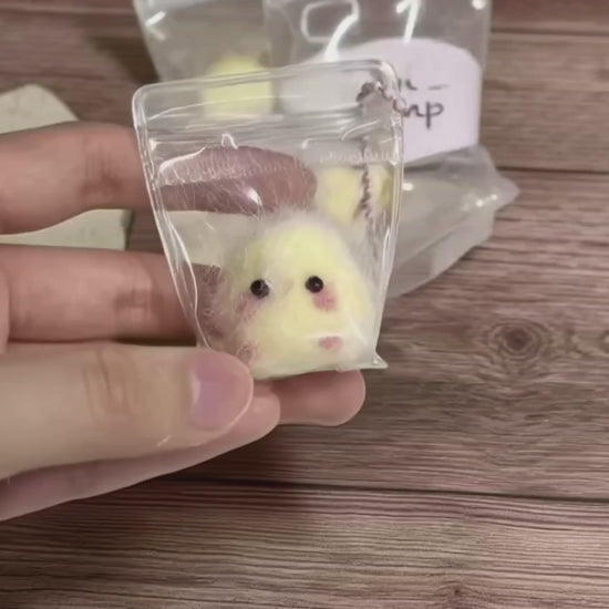 A short, close-up video of a hand holding a tiny, clear zip-lock bag. Inside is a "Mini Mochi Chick," a very small, fuzzy yellow plush with rosy cheeks. The person gently squeezes the bag, showing how soft and squishy the chick is. Other similar packages are visible in the background on a wooden table.