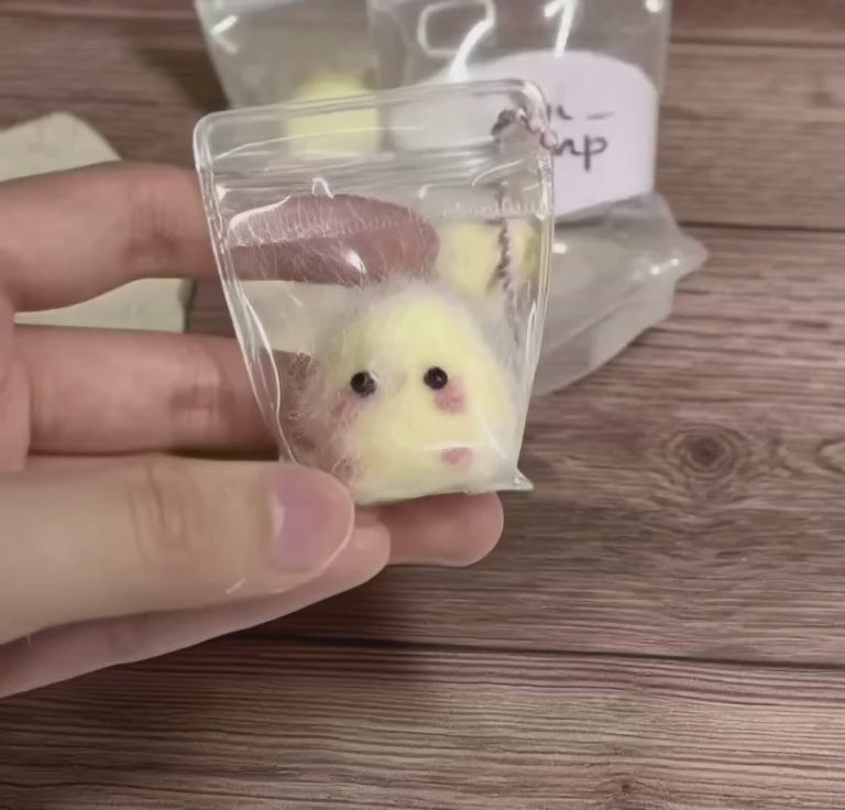 A short, close-up video of a hand holding a tiny, clear zip-lock bag. Inside is a "Mini Mochi Chick," a very small, fuzzy yellow plush with rosy cheeks. The person gently squeezes the bag, showing how soft and squishy the chick is. Other similar packages are visible in the background on a wooden table.